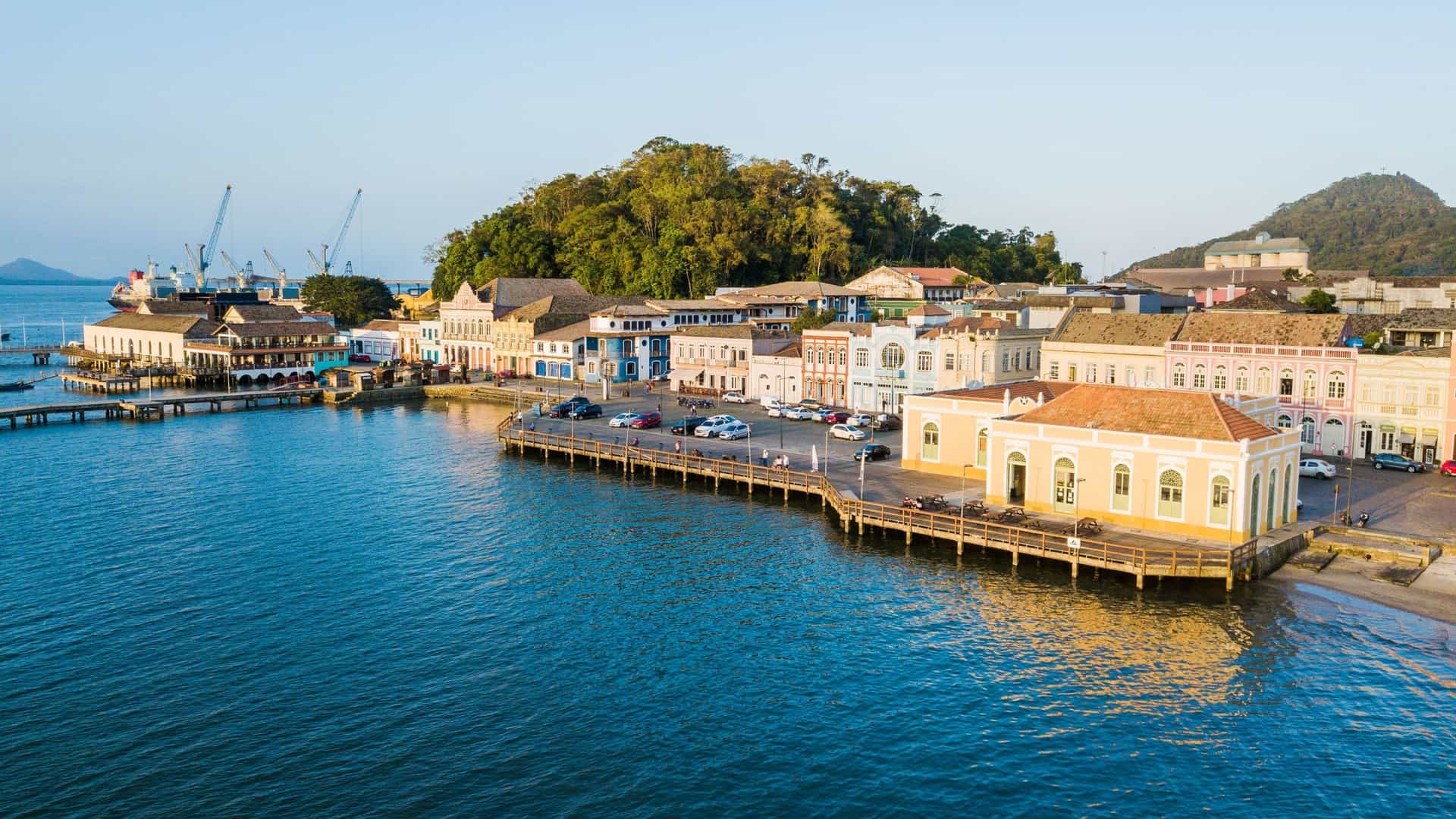 An aerial view of the historic São Francisco do Sul waterfront, featuring colorful colonial-era buildings lining a wooden pier beside the harbor.