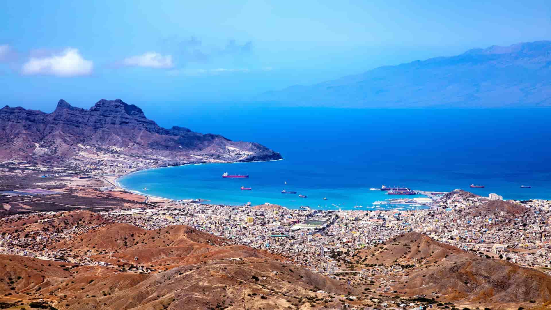 A breathtaking panoramic aerial view of the city of Mindelo on the island of São Vicente, Cape Verde, with its bustling harbor and town surrounded by rugged, volcanic hills.