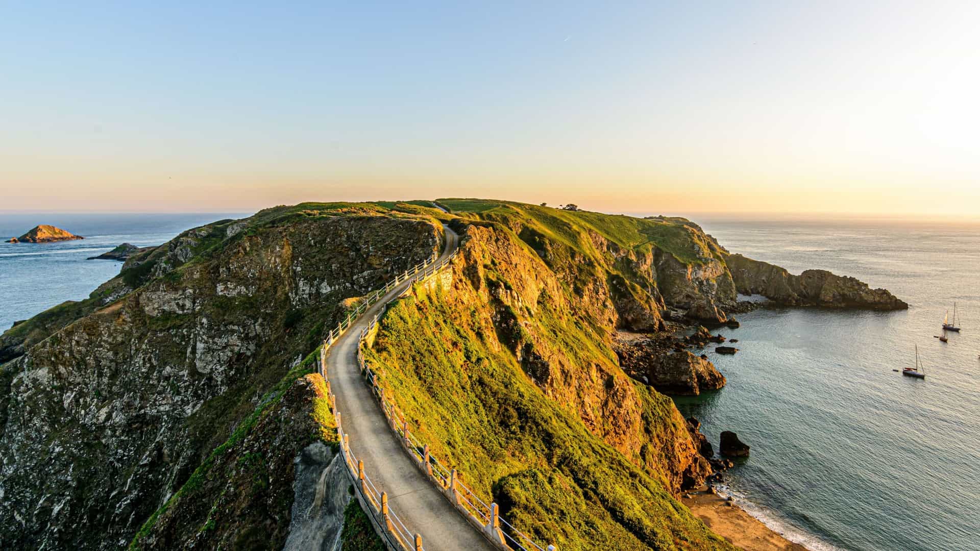 A stunning sunrise shot of La Coupée, a narrow, paved road on a high cliffside connecting two parts of the island of Sark, with the sea on both sides.