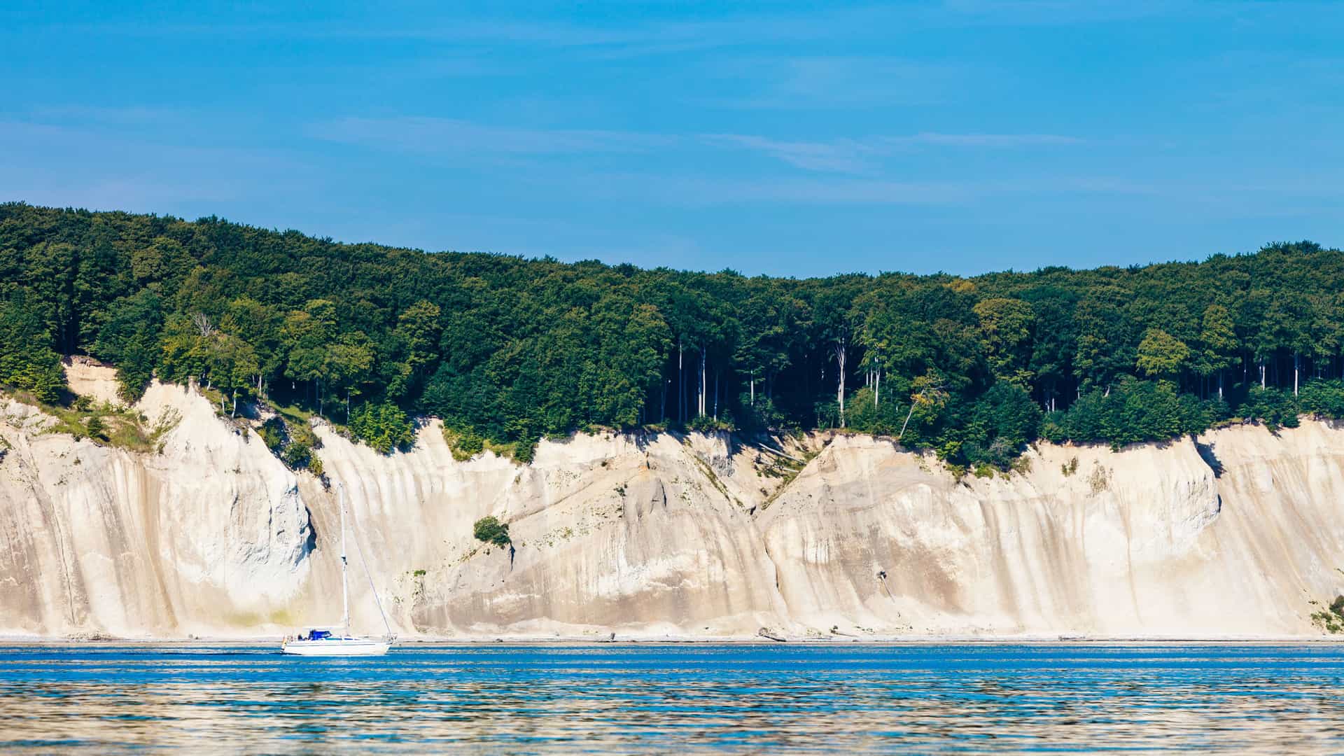 A serene view of a white sailboat sailing on the Baltic Sea, with a tall, white chalk cliff and a dense green forest in the background on the Isle of Rügen, Germany.