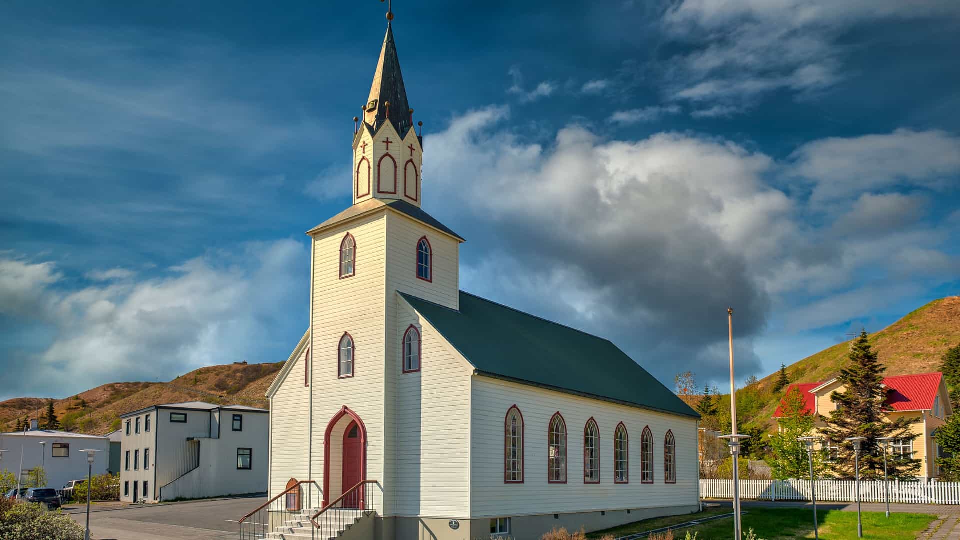 A wide-angle view of the historic Saudarkrokur Church in Iceland, a white wooden building with a red-framed door and steeple, set against a dramatic sky with hills in the background.