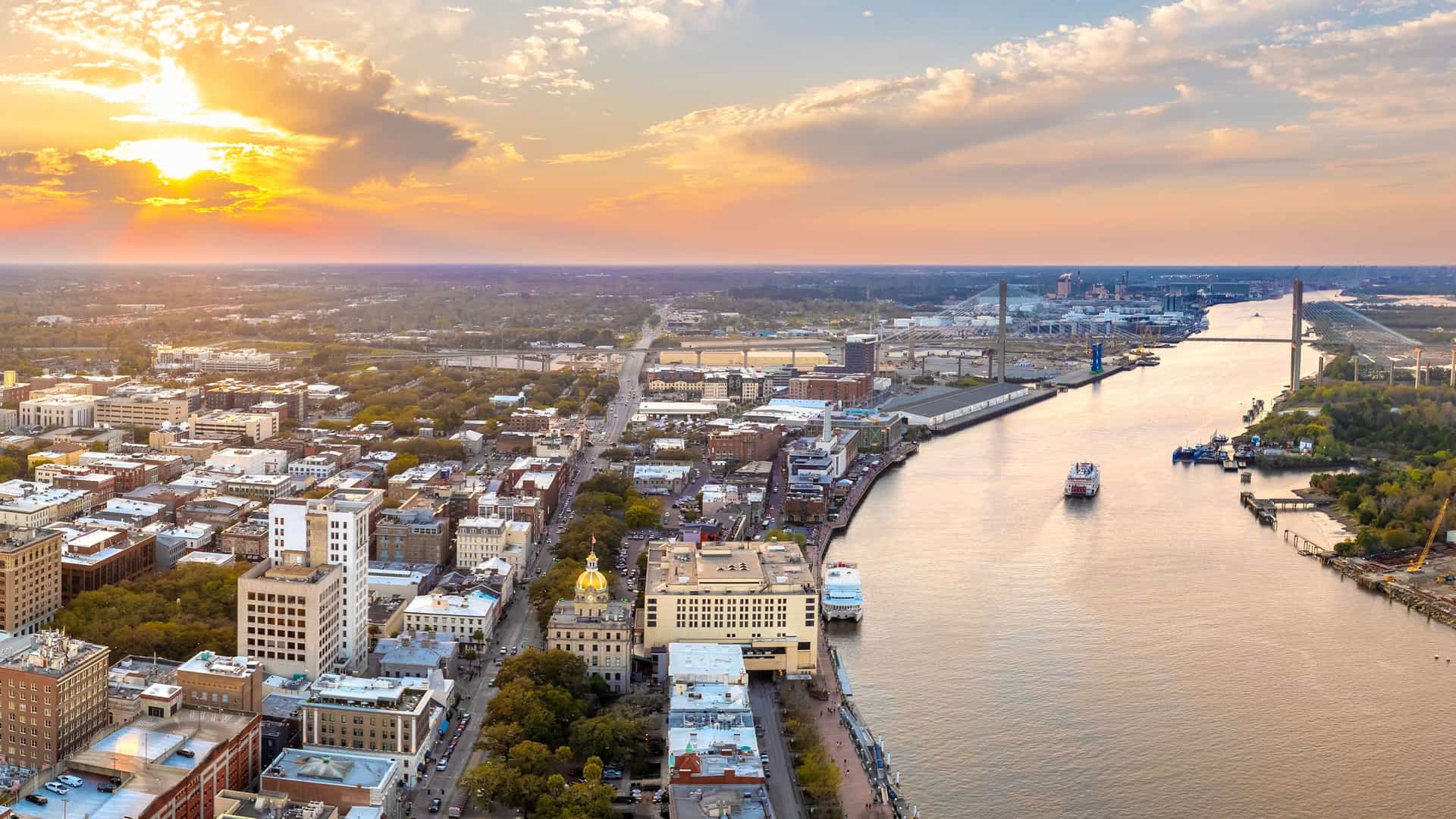 "An aerial sunset view of the Savannah, Georgia skyline, with the Savannah River flowing through the city's historic district and the Talmadge Memorial Bridge in the background. "