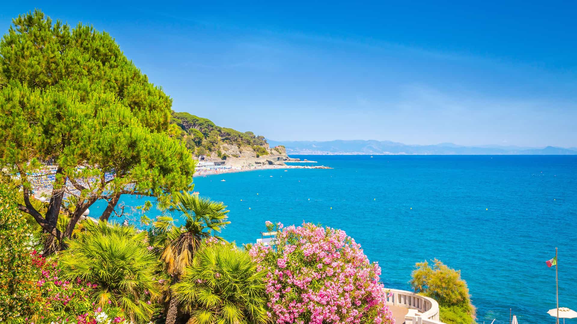 A breathtaking view of the scenic coastline in Savona, Italy, with lush green trees and vibrant pink flowers overlooking the deep blue Ligurian Sea.