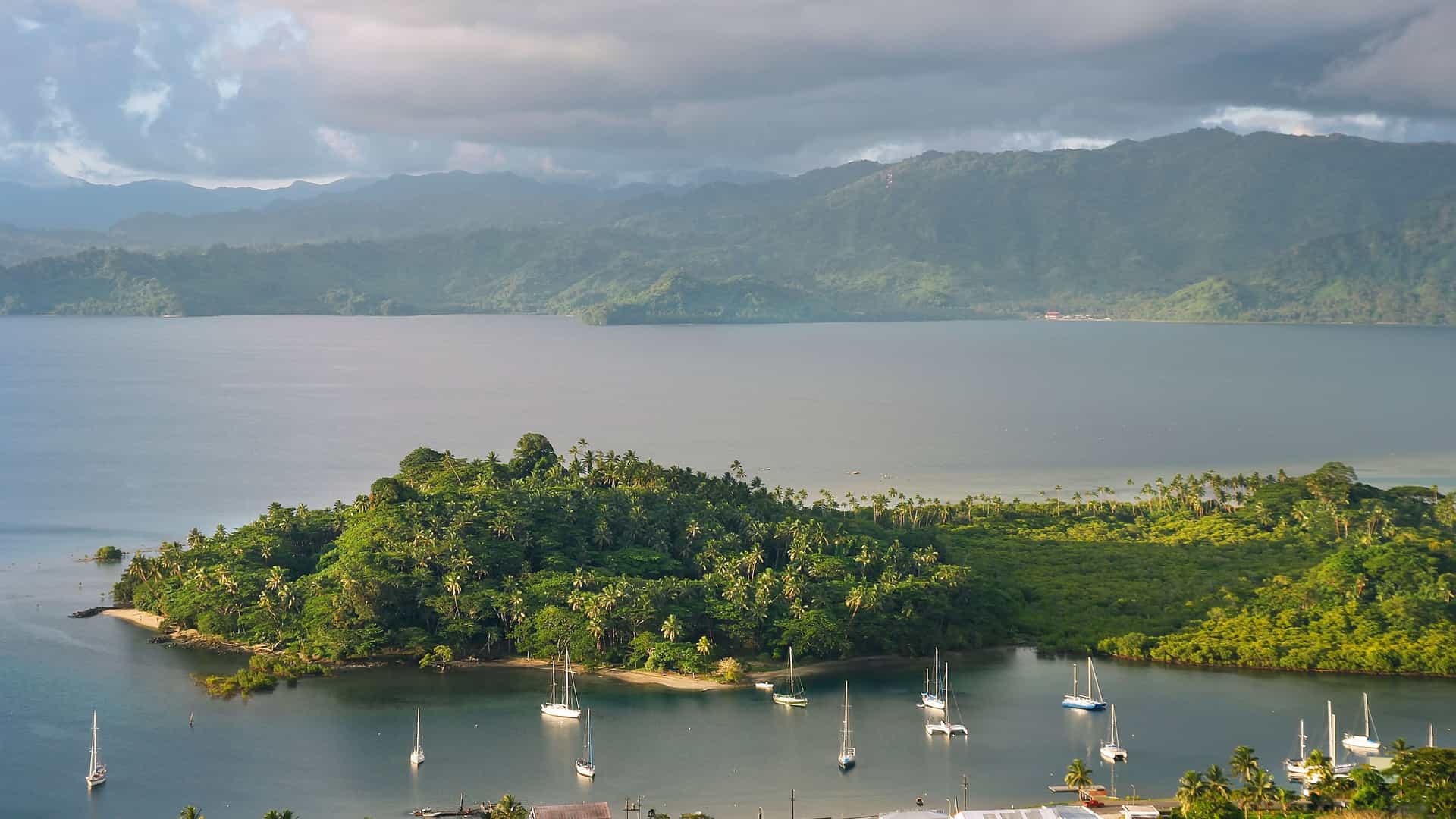 A panoramic view of Savusavu, Vanua Levu, Fiji, with a tranquil bay filled with sailboats, a lush green peninsula in the foreground, and mountainous terrain in the background.
