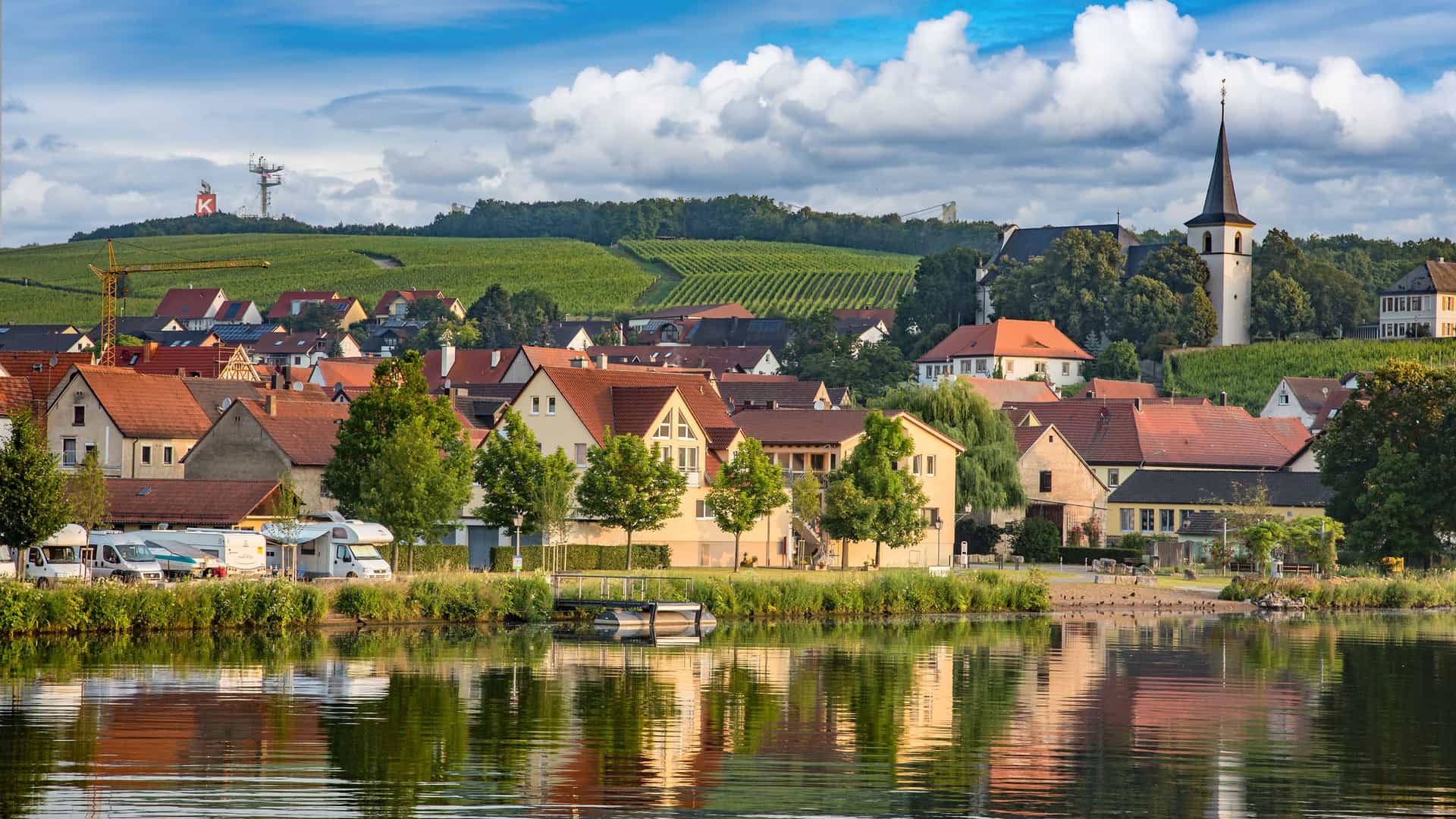 A panoramic view of the scenic town of Schweinfurt, Germany, with its historic buildings and a church steeple overlooking a calm lake, lush green vineyards, and rolling hills in the background.