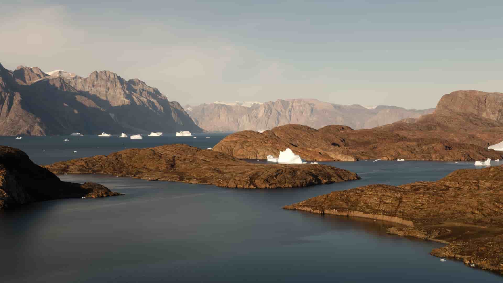 A breathtaking landscape shot of Scoresby Sund, Greenland, featuring floating icebergs and small brown islands surrounded by deep blue water and majestic mountains.