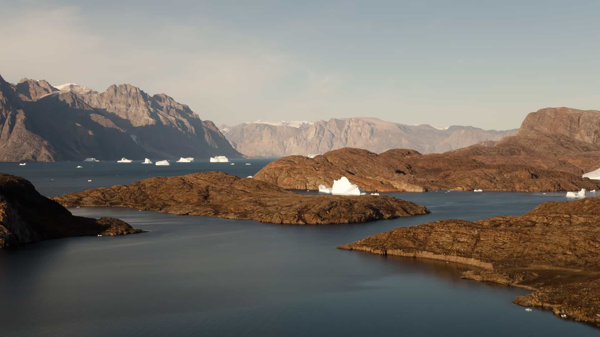 A breathtaking landscape shot of Scoresby Sund, Greenland, featuring floating icebergs and small brown islands surrounded by deep blue water and majestic mountains.