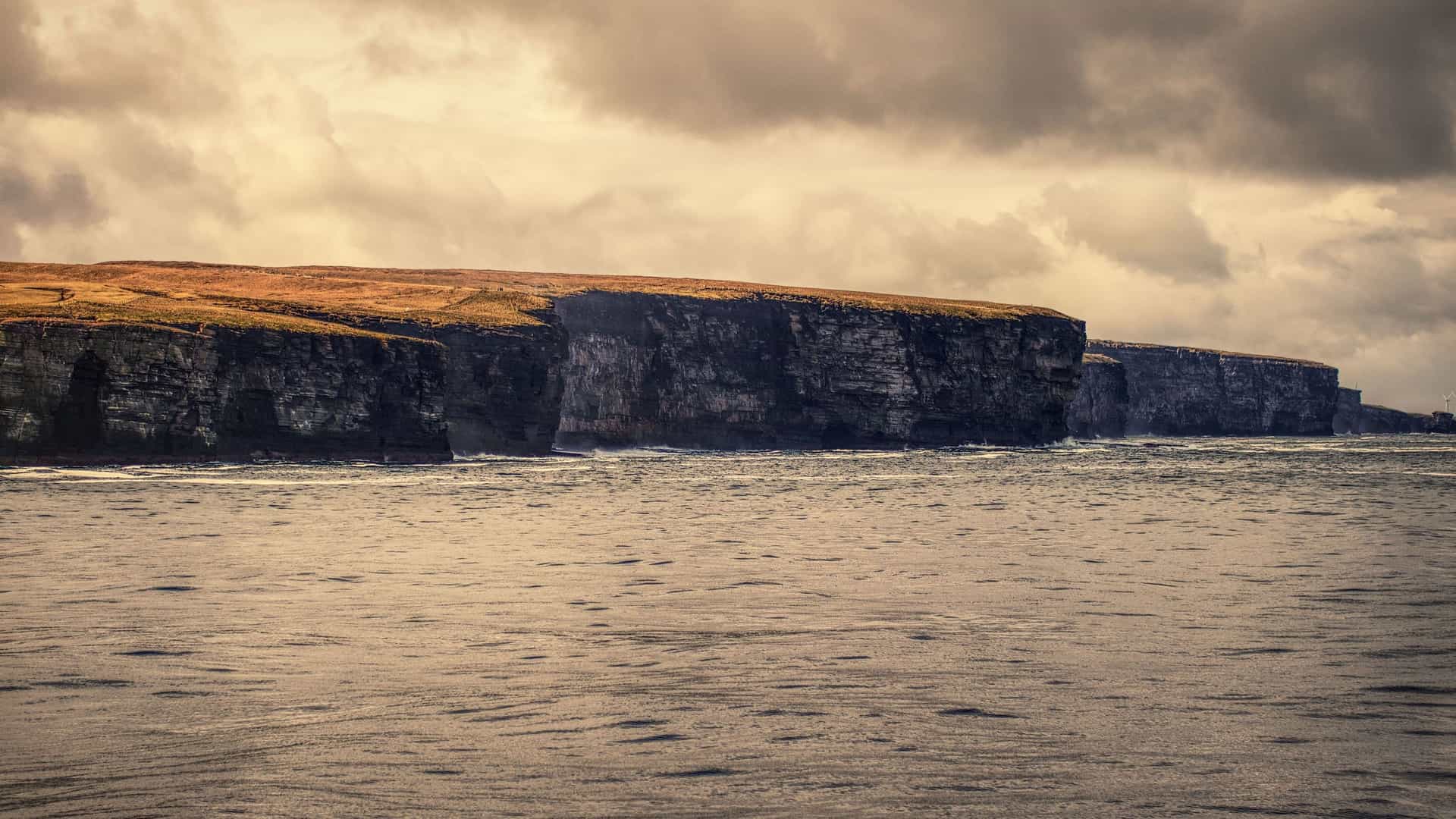A scenic view of the dramatic, high sea cliffs on the coast of Scrabster, Scotland, with the vast, dark waters of the Pentland Firth in the foreground and a moody sky above.