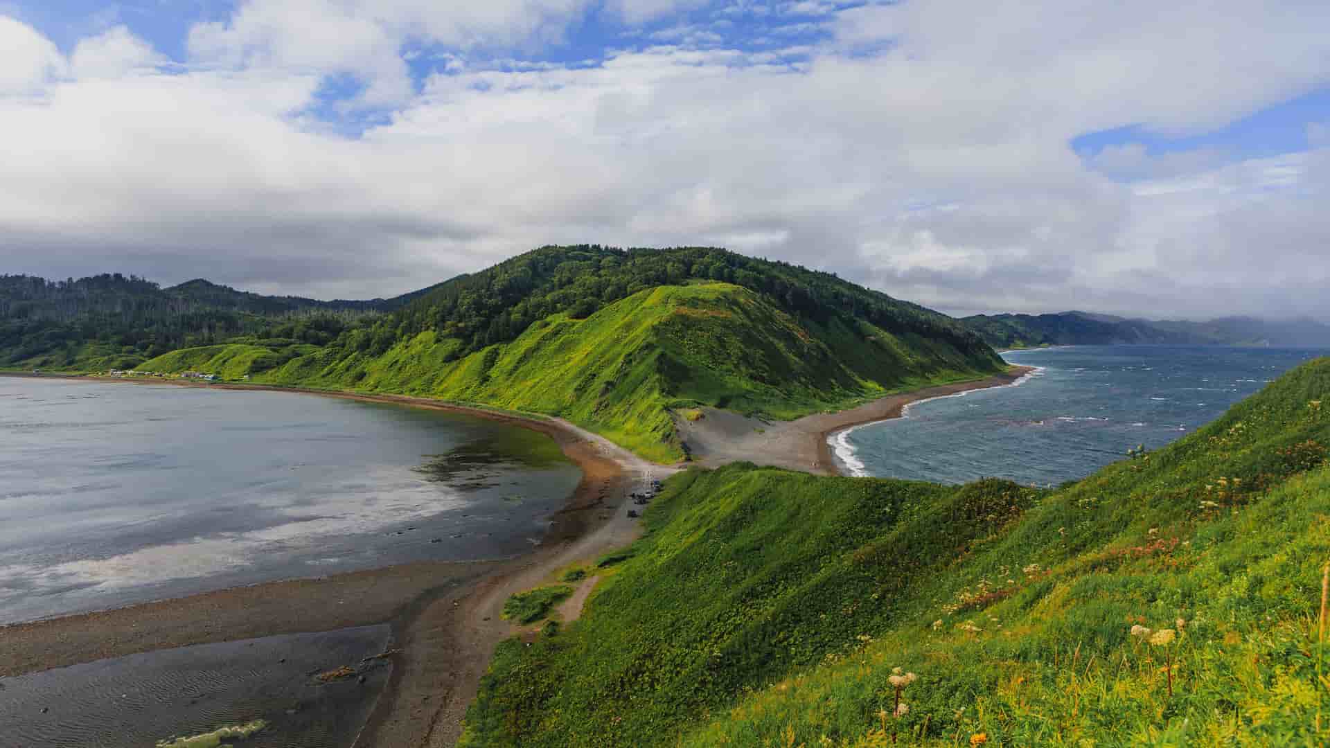 A stunning wide-angle view of the scenic coastline of the Sea of Okhotsk in Russia, with a narrow land bridge separating the tranquil bay from the deep blue sea, all surrounded by lush green hills.