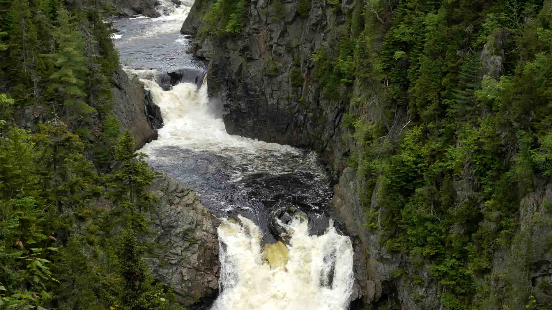 A dramatic view of the Sainte-Anne-du-Nord River cascading through a deep, rocky gorge surrounded by dense green forest near Sept-Îles, Quebec.