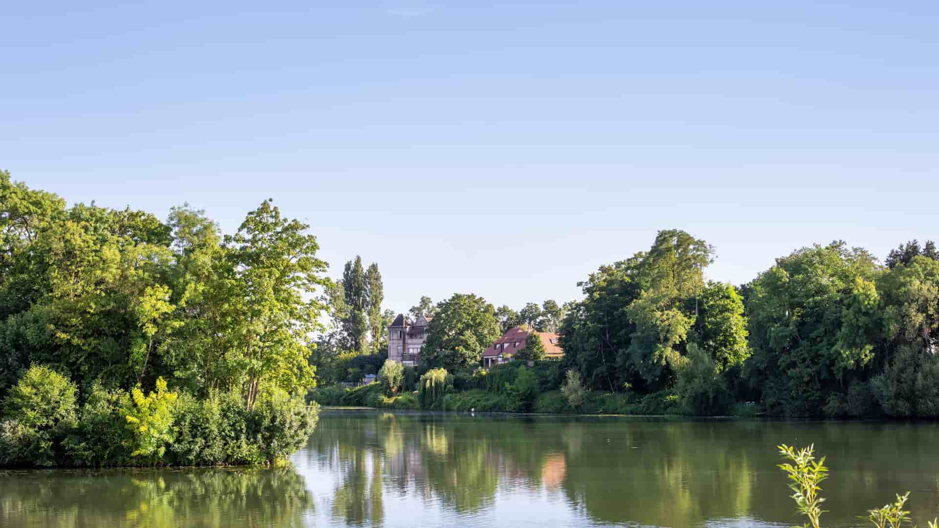 A tranquil wide-angle view of the Saône River in Seurre, France, with lush green trees lining the shore and historic houses reflected in the calm water under a clear blue sky.