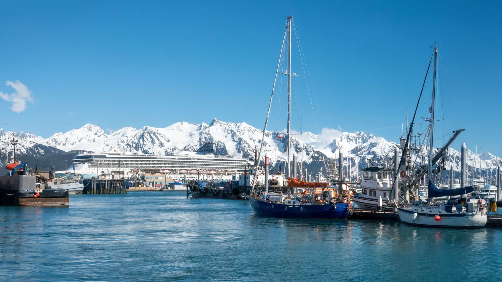 "A large cruise ship and several sailboats docked in the harbor of Seward, Alaska, with the Chugach Mountains' snow-covered peaks visible in the distance. "
