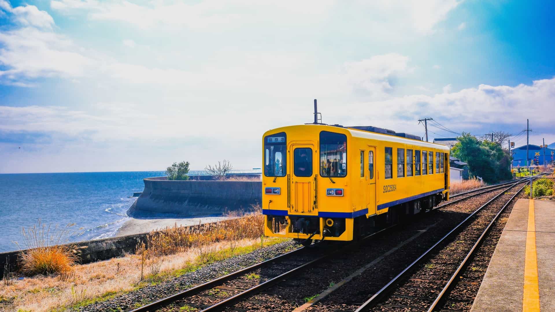 A bright yellow train on the coastal rail line in Shimabara, Japan, running beside the blue ocean under a sunny sky.