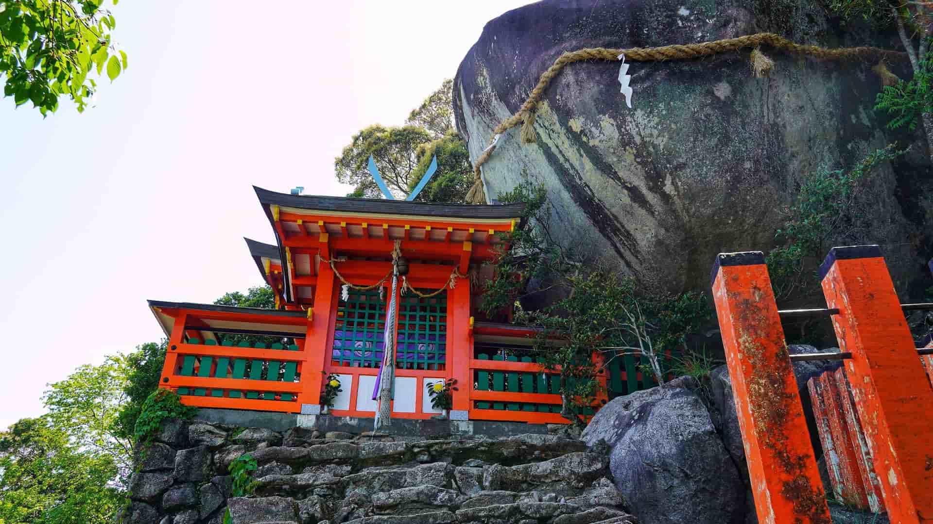 A vibrant red Shinto shrine with an orange roof and a bright green fence, built at the base of a massive, sacred rock, in Shingu, Wakayama, Japan.