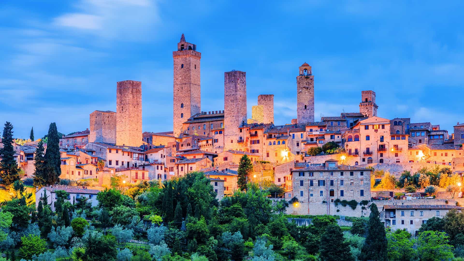 "A breathtaking twilight view of the medieval city of Siena, Italy, with its dramatic towers and historic buildings illuminated against the clear blue evening sky. "