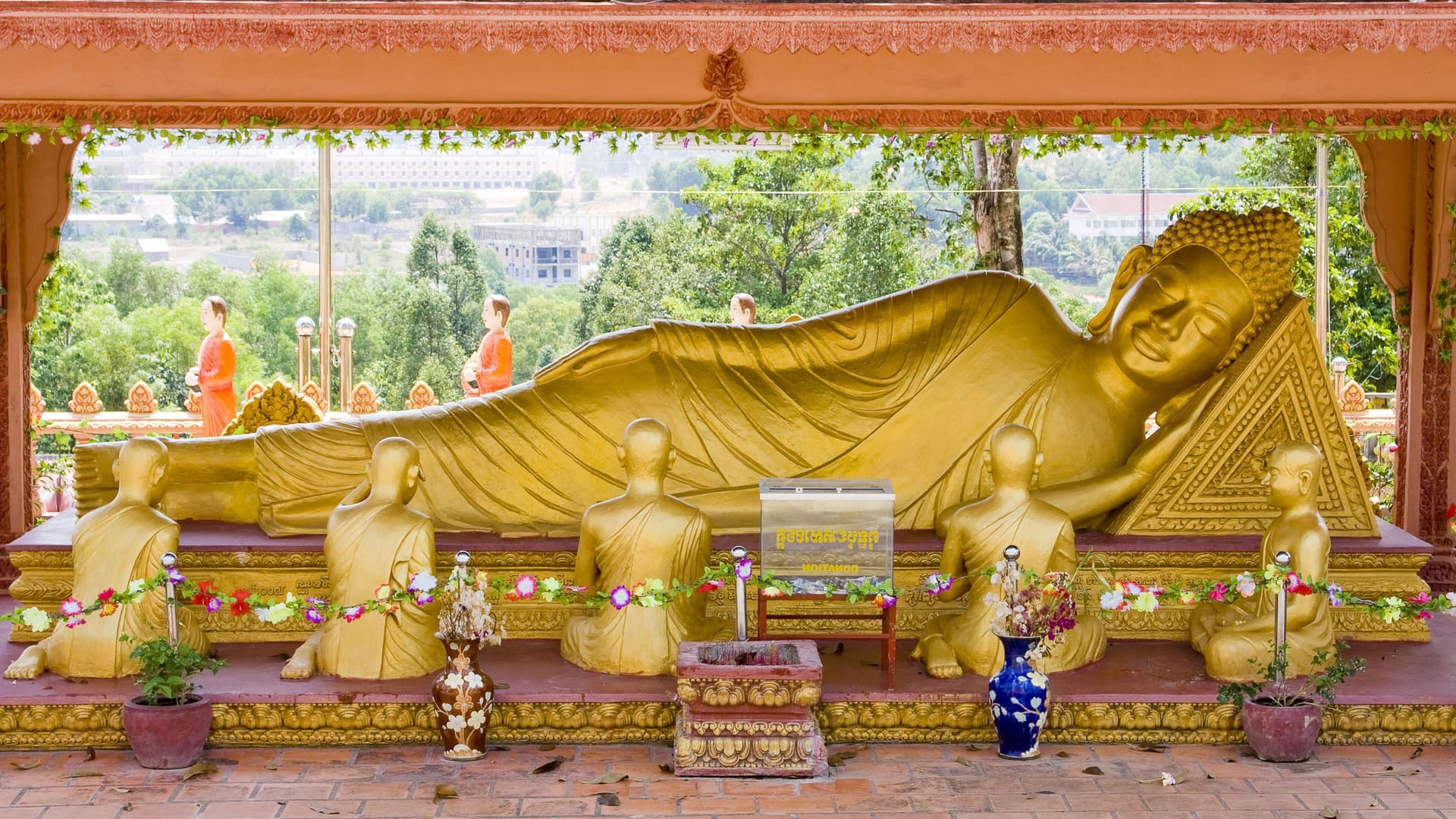 A golden reclining Buddha statue, adorned with flowers and surrounded by smaller statues of monks, is housed in a pagoda with a view of the countryside near Sihanoukville.