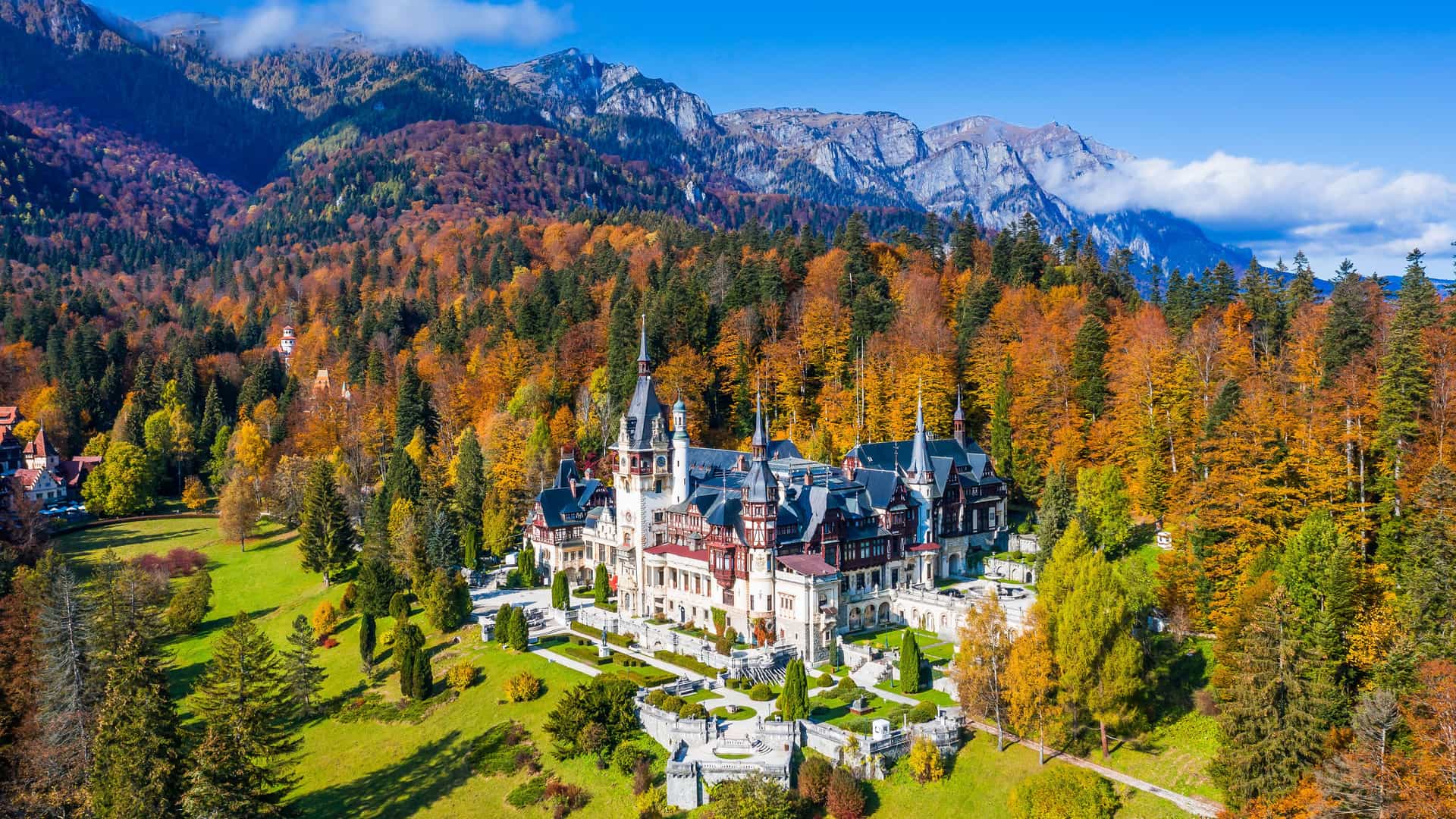 A high-angle view of the beautiful Peleș Castle in Sinaia, Romania, a neo-renaissance building nestled in the Carpathian Mountains, surrounded by lush autumn foliage and gardens.