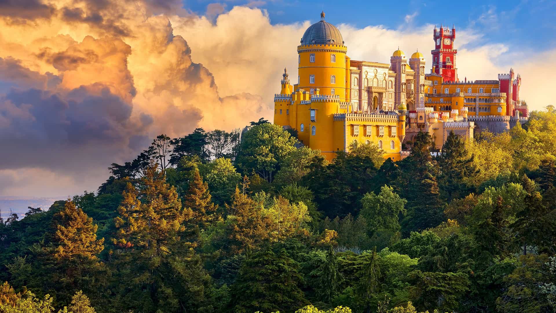 A high-angle view of the colorful Pena Palace, a romanticist castle with red and yellow towers, perched on a mountaintop in the Sintra Mountains of Portugal.