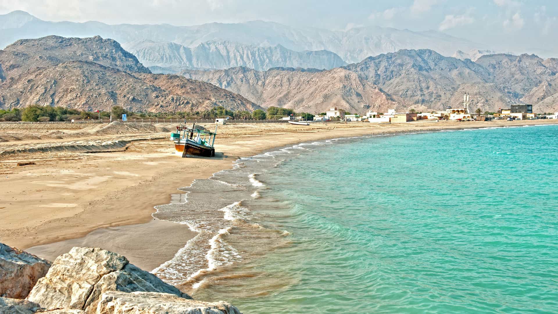 A serene scenic view of Sir Bani Yas Island, with a small traditional boat on a sandy beach and turquoise water, framed by rocky mountains in the background.