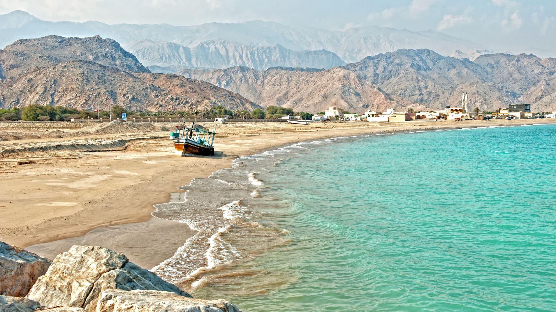 A serene scenic view of Sir Bani Yas Island, with a small traditional boat on a sandy beach and turquoise water, framed by rocky mountains in the background.