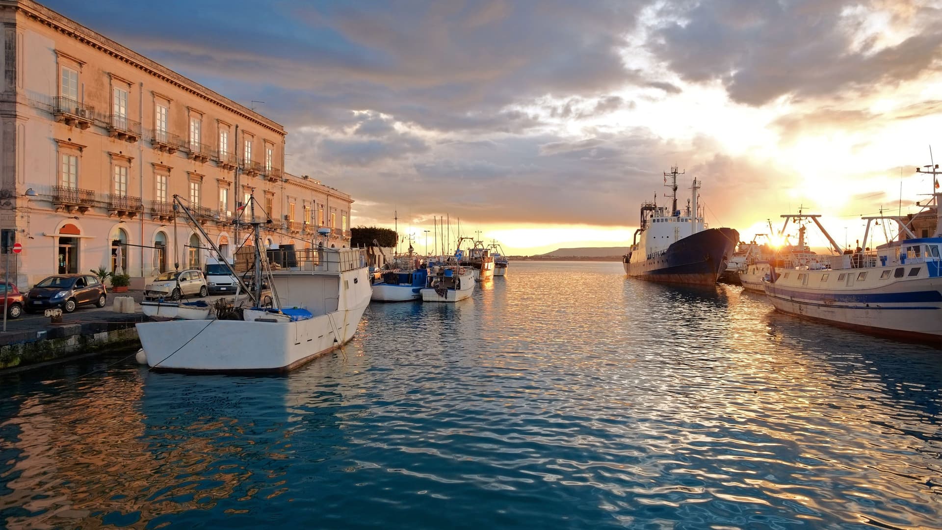 A picturesque sunset shot of the harbor in Siracusa, Sicily, with a row of historic buildings along the waterfront and fishing boats moored in the glowing water.