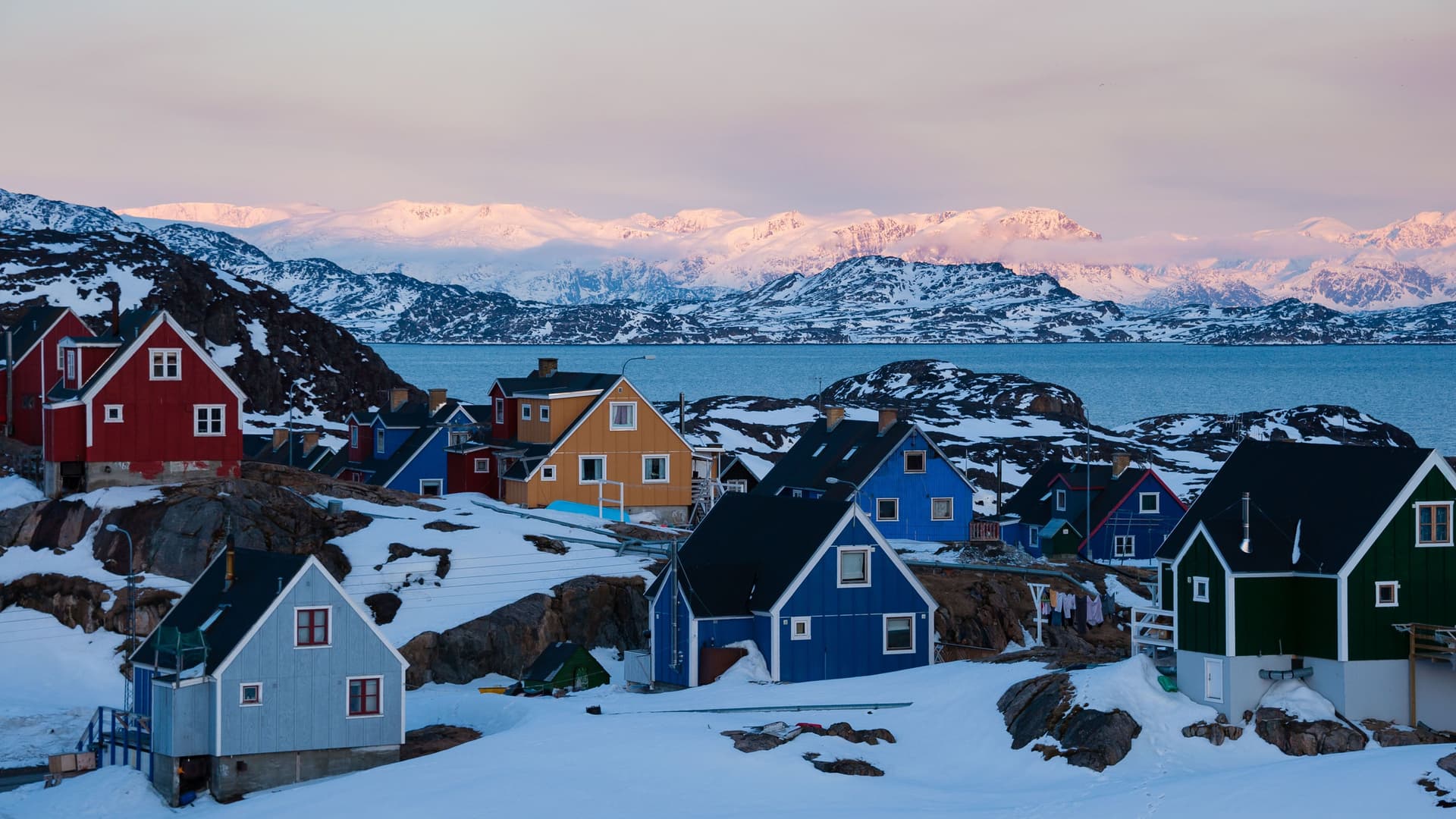 A vibrant shot of colorful wooden houses in the snow-covered town of Sisimiut, Greenland, with a view of the icy sea and snow-capped mountains at sunrise.