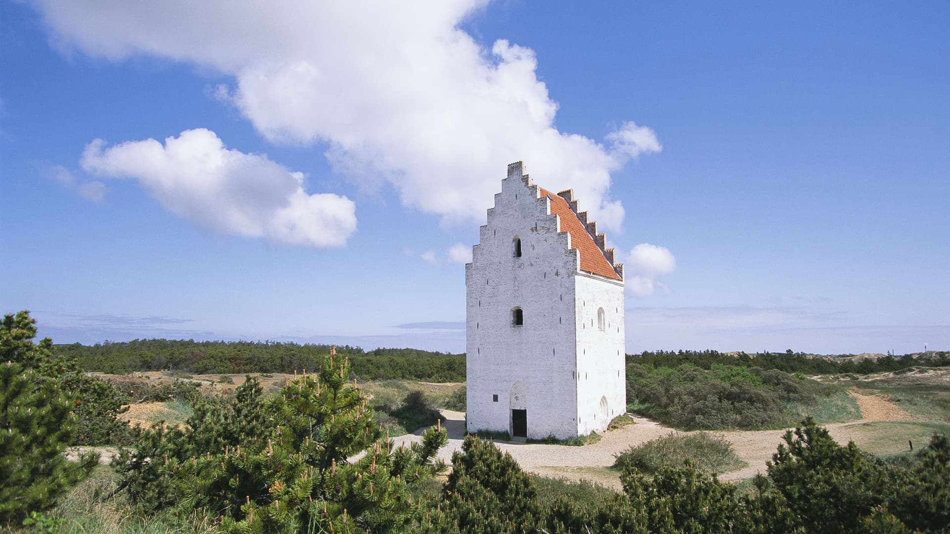 A striking image of Den Tilsandede Kirke, or the Buried Church, a historic white church with a red roof partially submerged in sand dunes near Skagen, Denmark.
