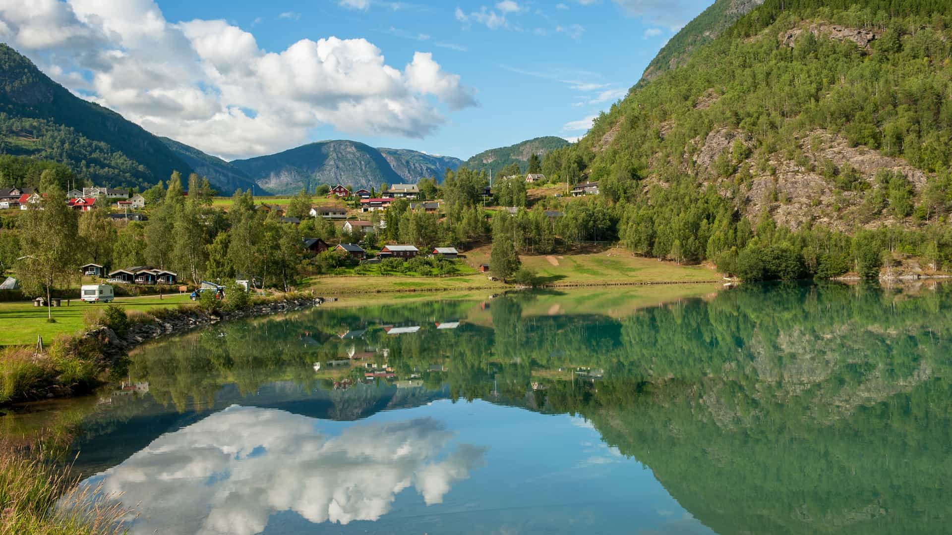 A serene landscape shot of Skjolden, Norway, with a small village nestled in a lush green valley between mountains, reflected beautifully in a calm lake.