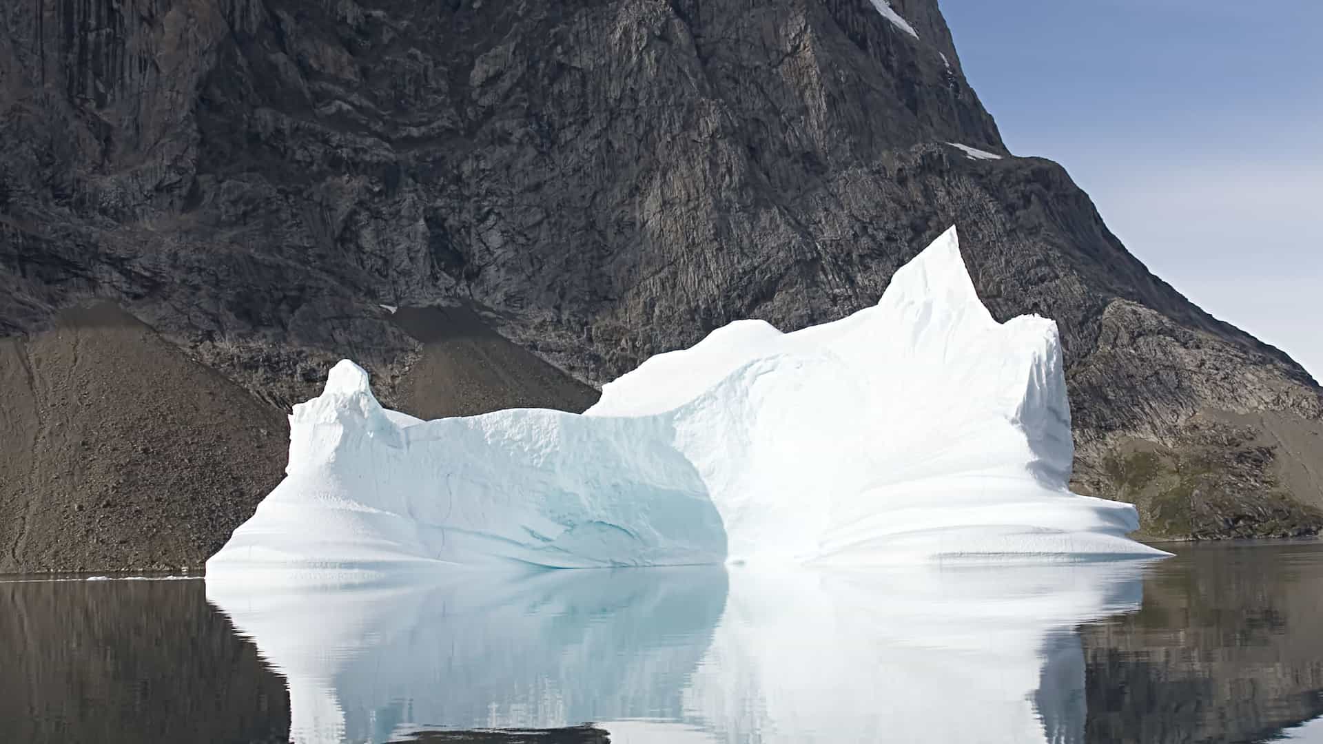 A breathtaking view of a massive, white iceberg floating in a calm body of water in the Skjoldungen Island Fjord in Greenland, with a towering, rocky mountain in the background.