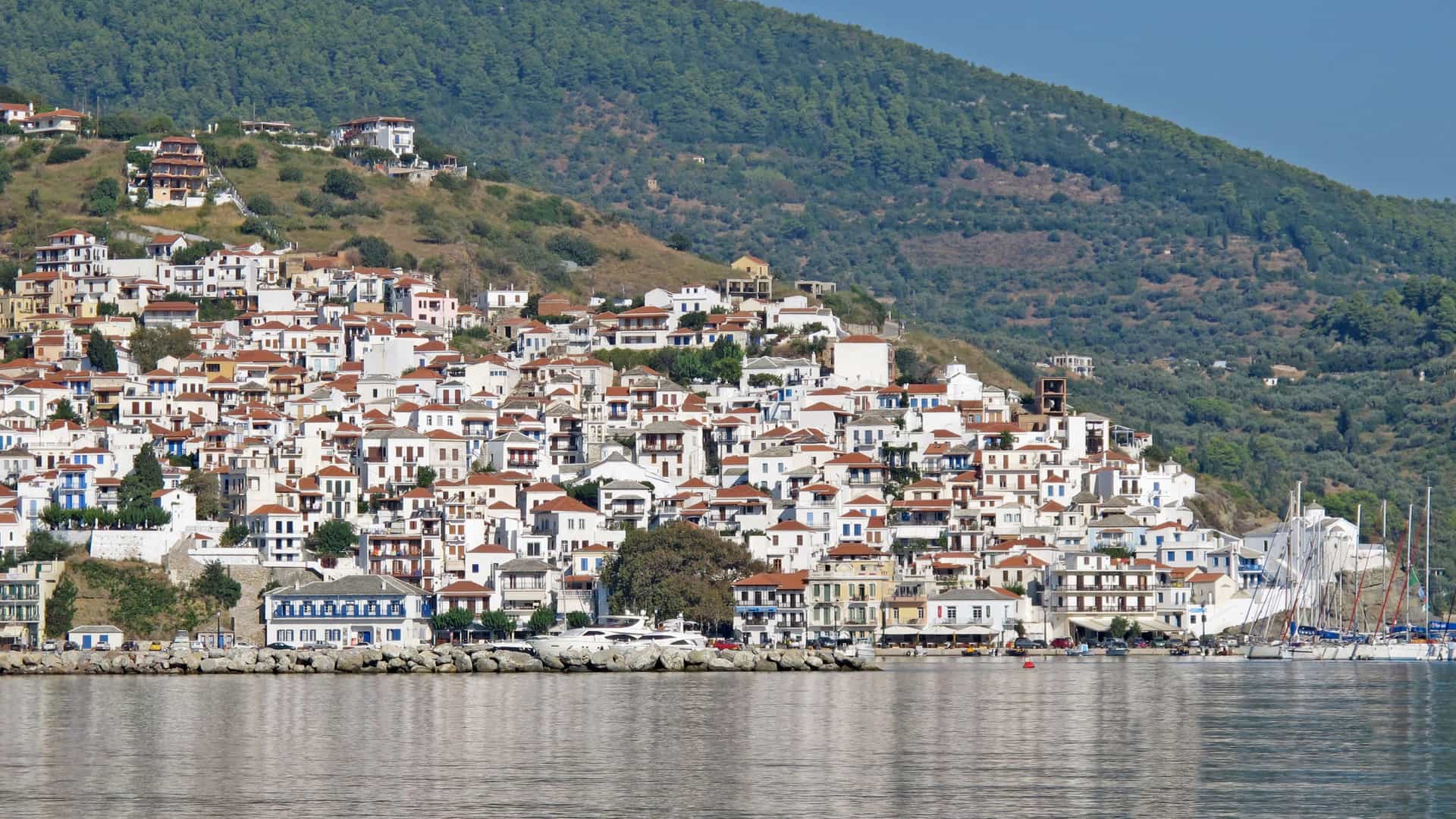 A scenic view of Skopelos Town on Skopelos Island, Greece, with its white buildings and red-tiled roofs cascading down a lush green hillside to the tranquil harbor.