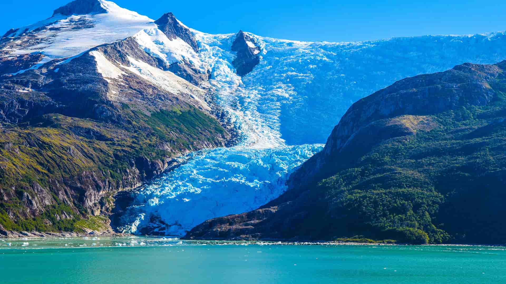 A panoramic view of a massive blue glacier flowing down a mountain and into the turquoise waters of the Smyth Channel in Chile.