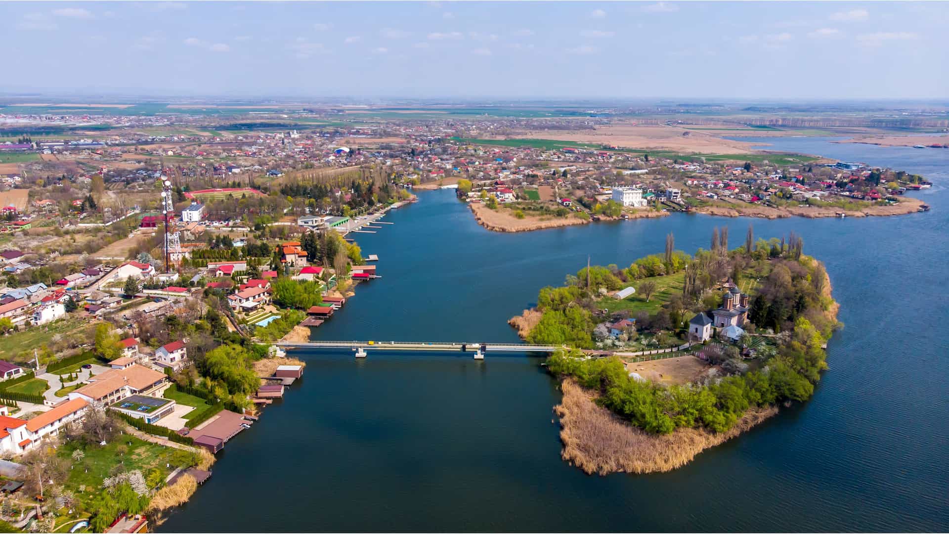 An aerial view of the scenic Snagov Lake in Romania, with a bridge connecting the mainland to a small island containing the historic Snagov Monastery.