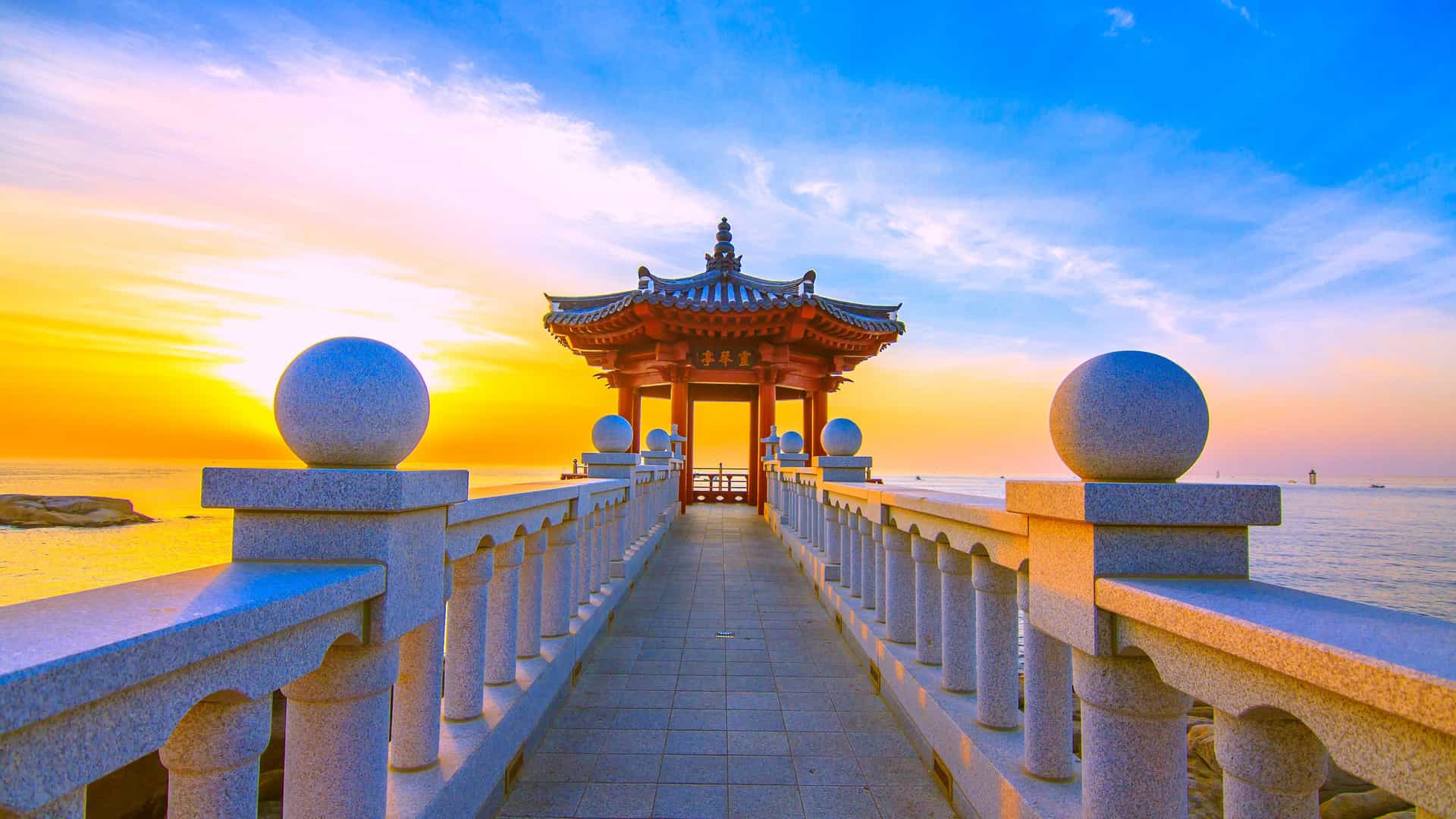 A scenic view of a traditional Korean pavilion at the end of a stone walkway, overlooking the East Sea in Sokcho, South Korea, with a stunning sunset in the background.