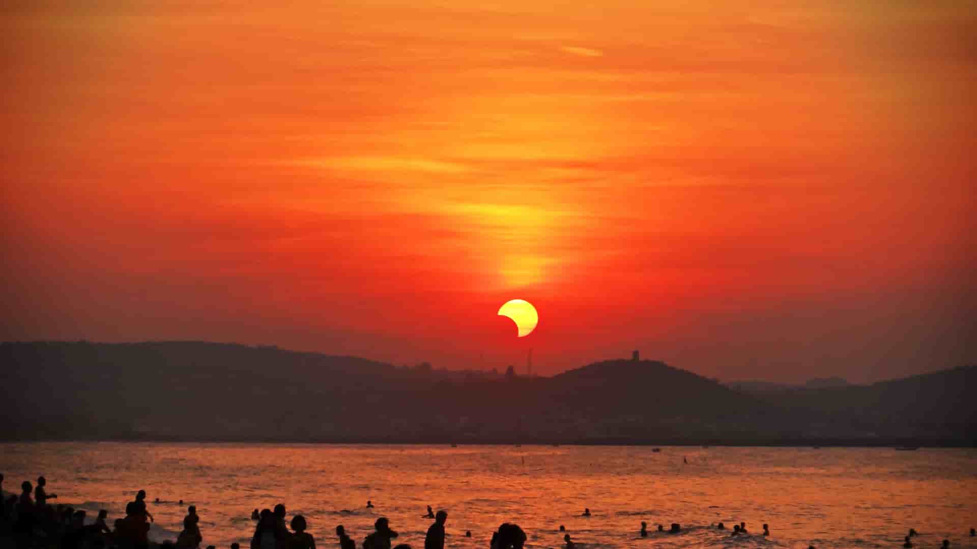 A crowd of people on a beach and in the water, watching a partial solar eclipse as the crescent-shaped sun sets over the ocean and distant hills.