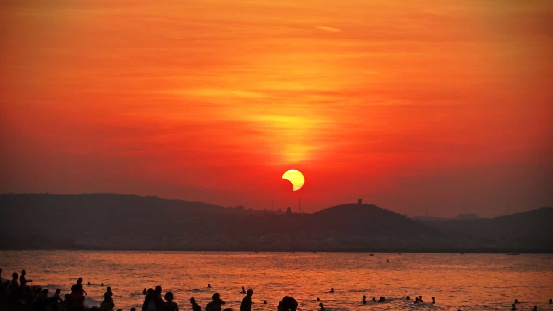 A crowd of people on a beach and in the water, watching a partial solar eclipse as the crescent-shaped sun sets over the ocean and distant hills.