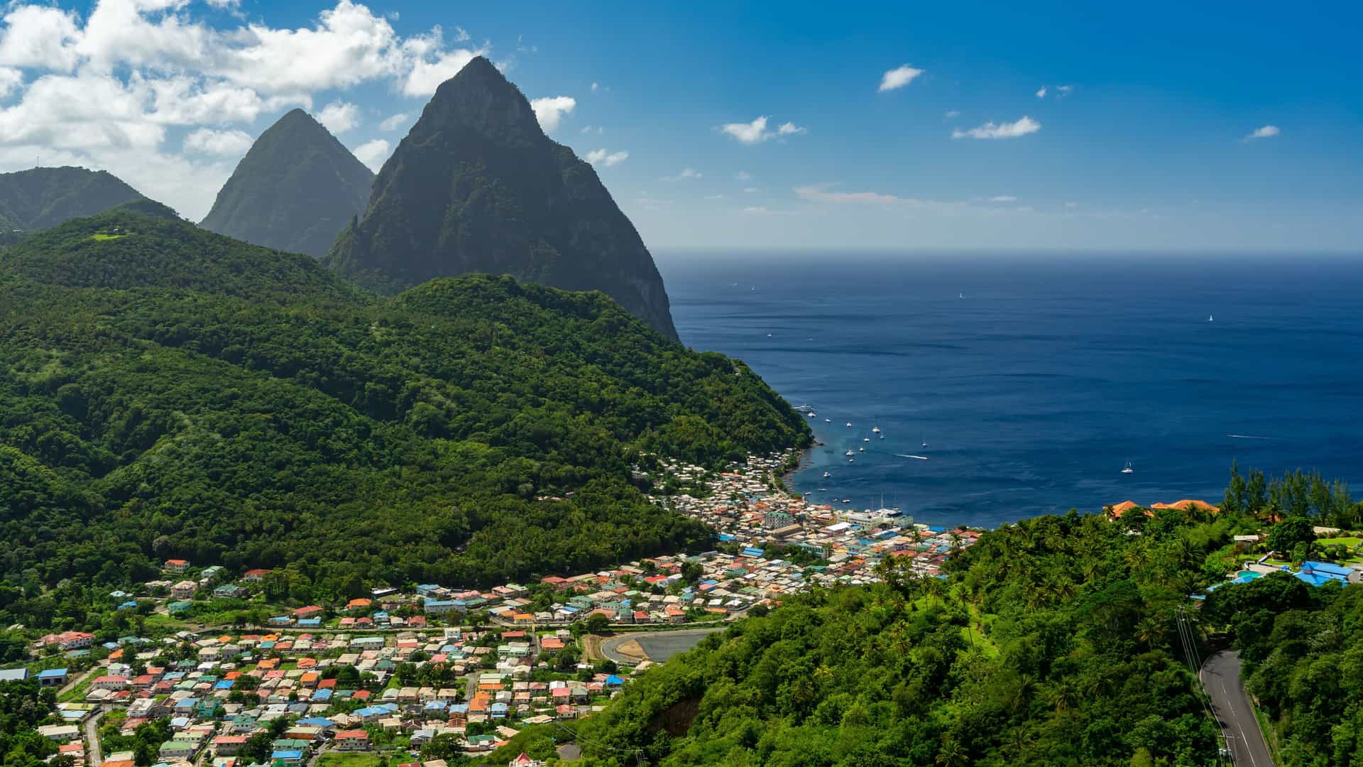 "A scenic view of the coastal town of Soufrière, St. Lucia, with its colorful houses nestled beneath the famous twin volcanic peaks, the Pitons, and the Caribbean Sea. "
