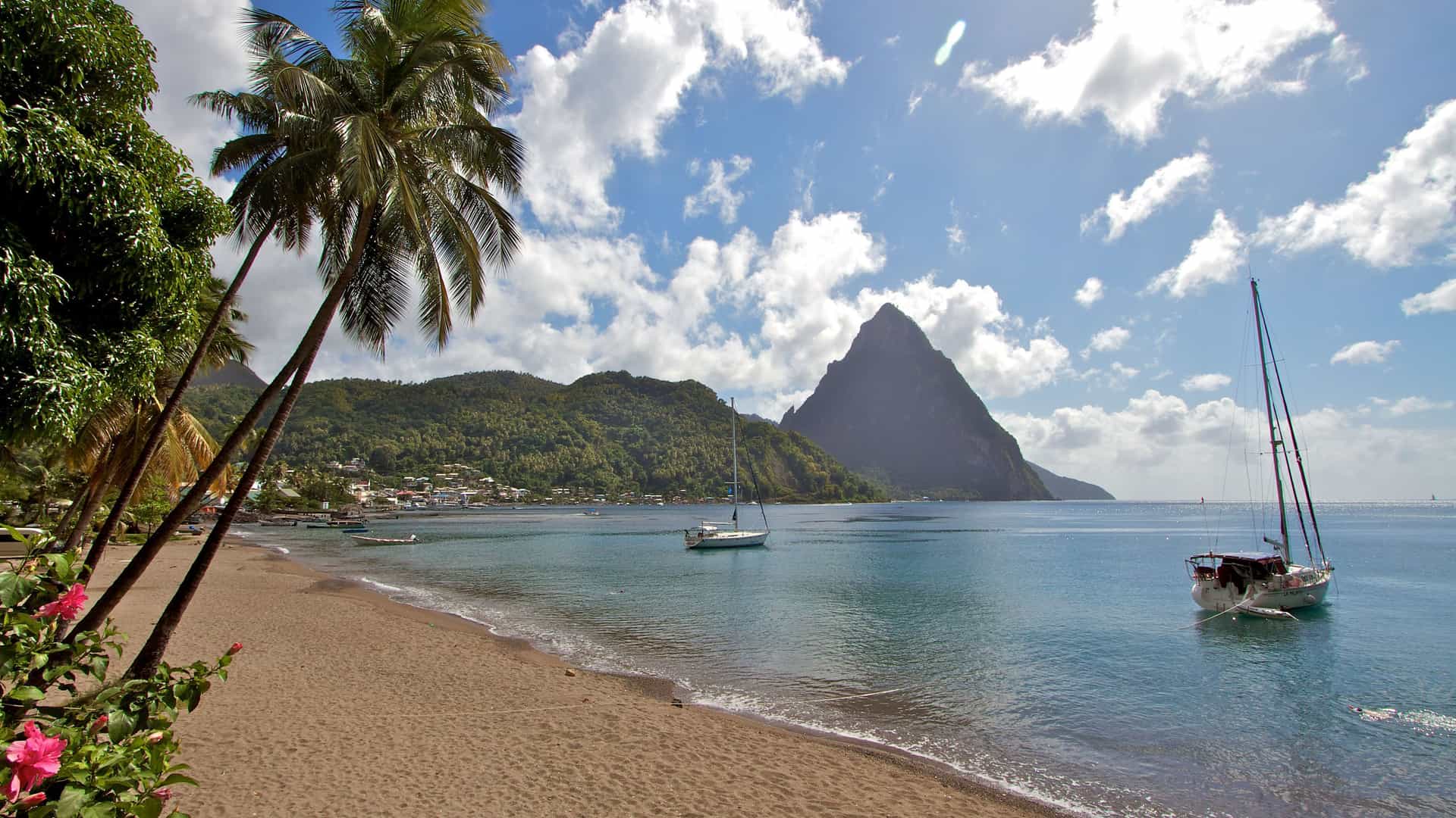 A tropical beach with golden sand, swaying palm trees, and two sailboats anchored in the bay in front of the lush green mountains of Soufrière, St. Lucia.