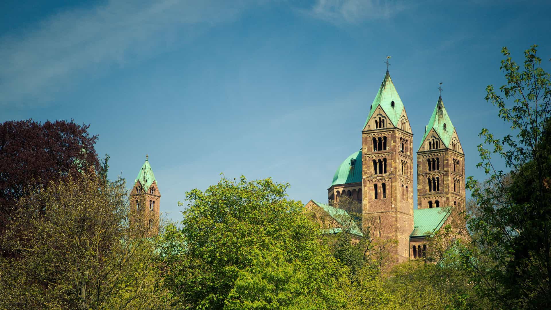 A beautiful view of the Speyer Cathedral with its iconic twin green-roofed towers peeking above the lush green trees against a clear blue sky in Speyer, Germany.