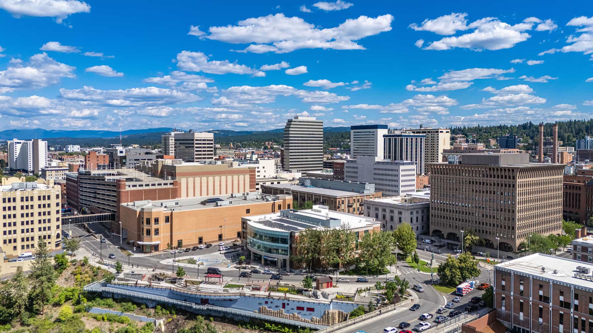 "An aerial view of the Spokane, Washington skyline, with its downtown buildings and the surrounding hilly landscape under a bright, cloud-filled blue sky. "