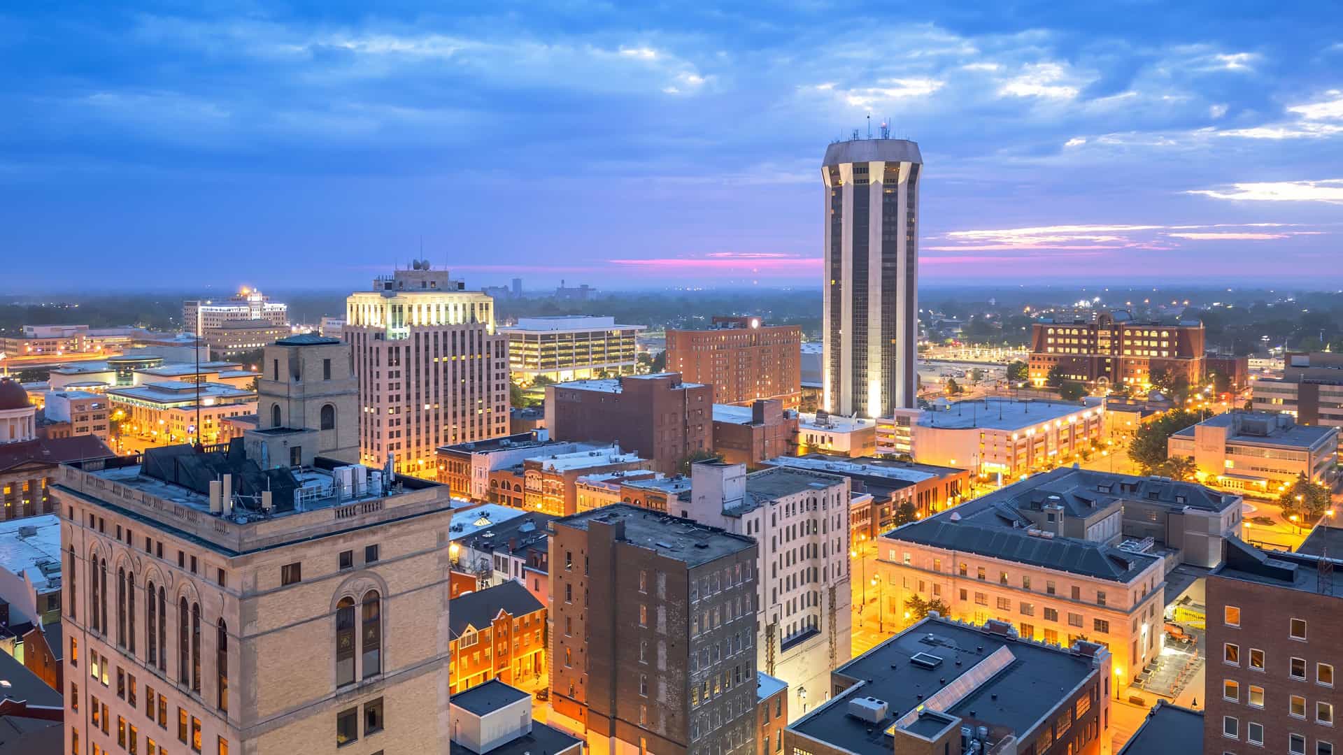 "An aerial twilight view of the Springfield, Illinois, skyline with its historic buildings and modern high-rises, illuminated against a dramatic pink and blue sky. "