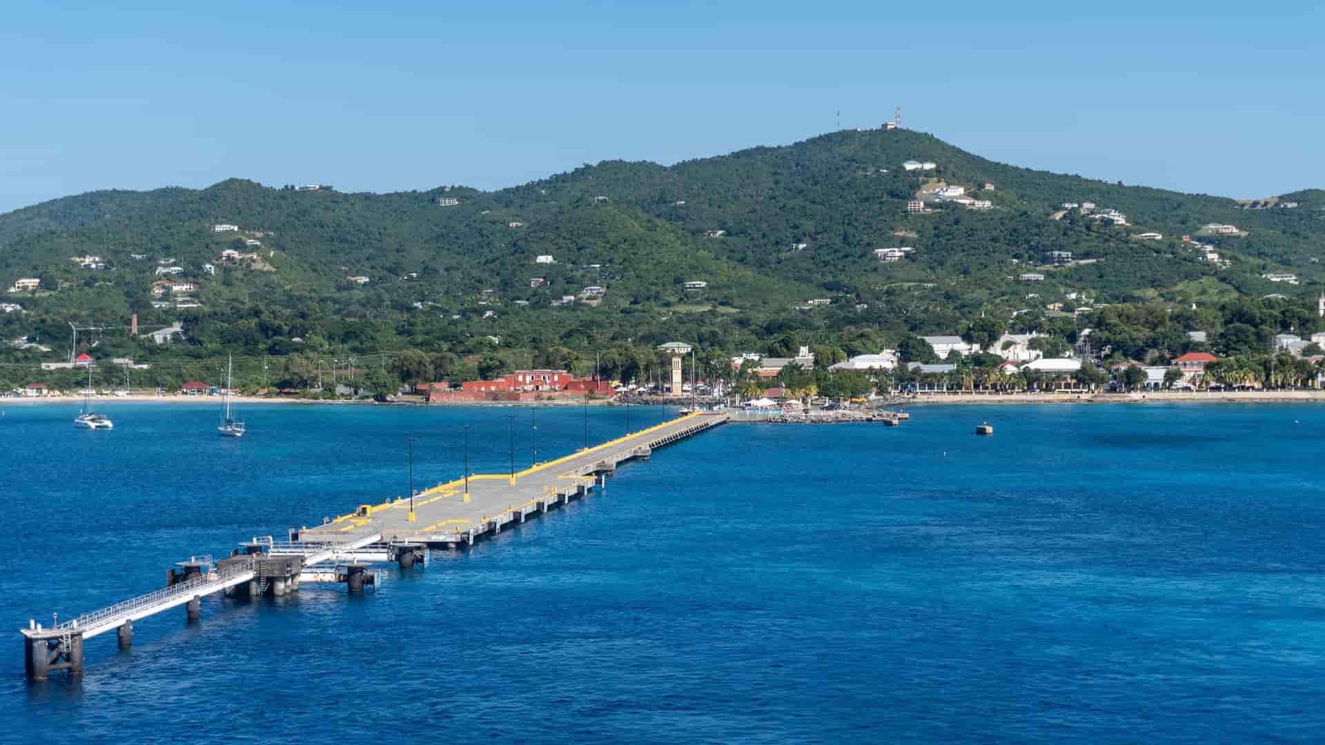 A panoramic view of a long, gray pier extending into the calm turquoise waters of St. Croix, US Virgin Islands, with the town and a lush green mountain in the background.