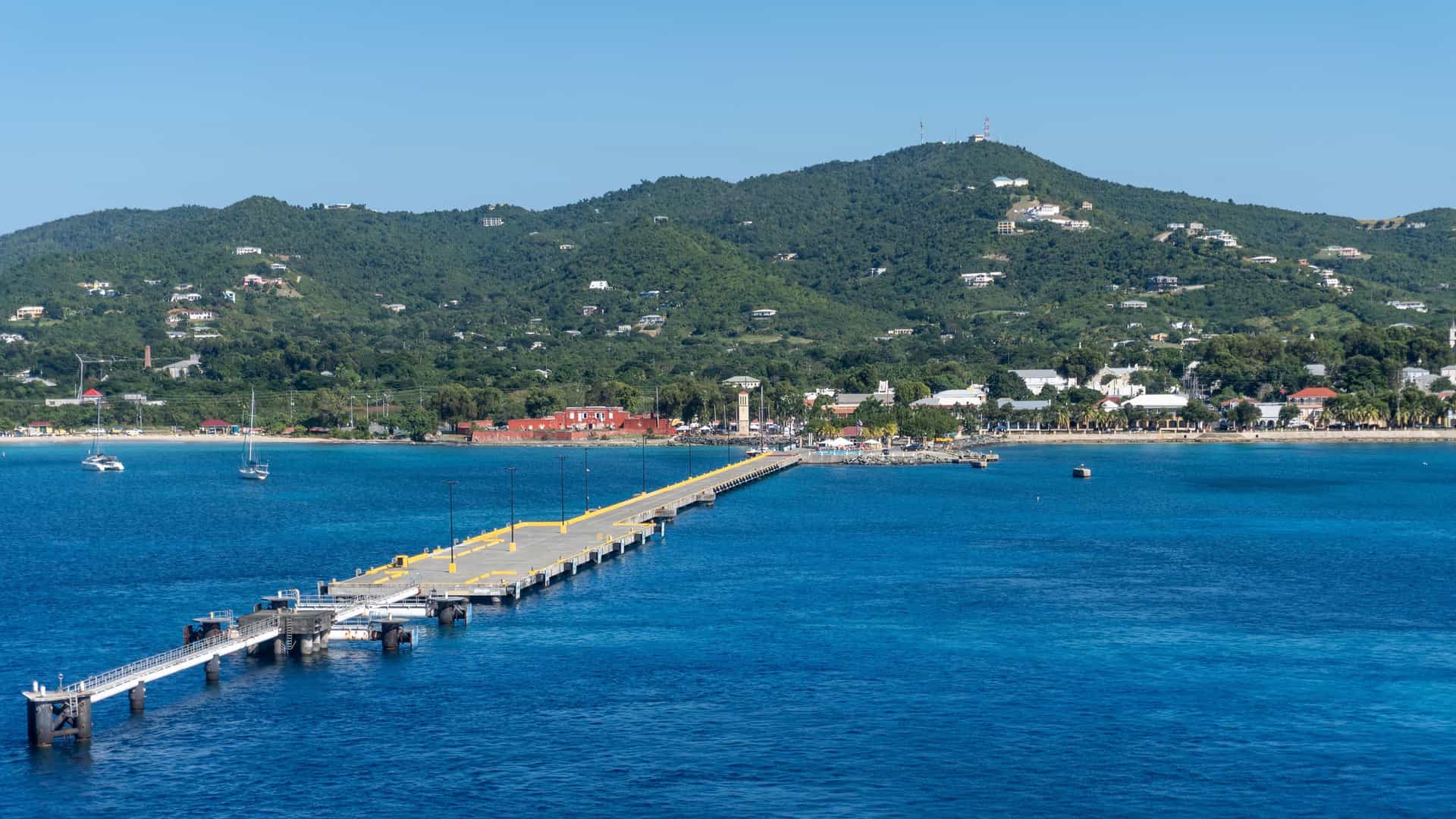 A panoramic view of a long, gray pier extending into the calm turquoise waters of St. Croix, US Virgin Islands, with the town and a lush green mountain in the background.