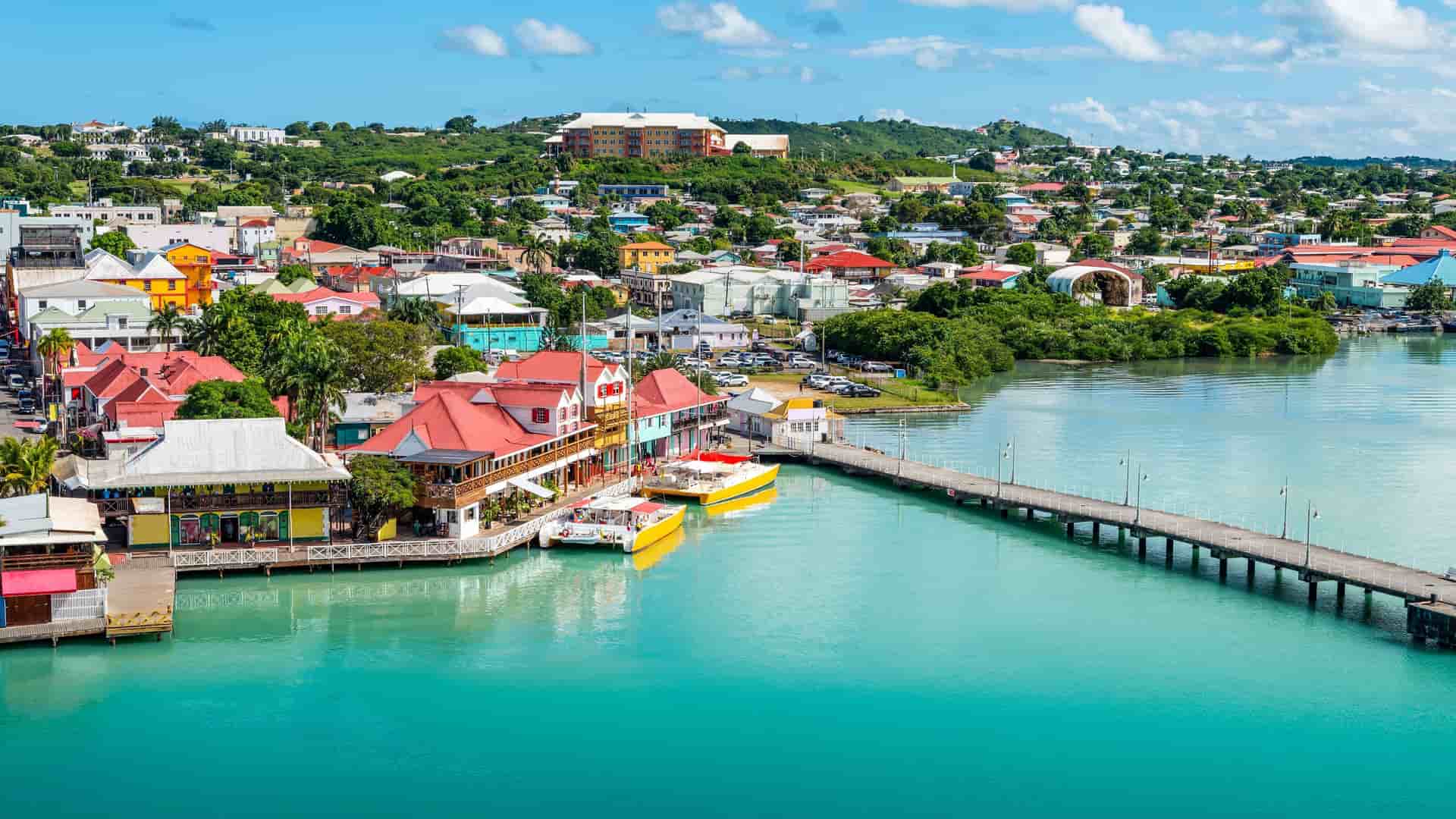 An aerial view of the colorful waterfront of St. John's, Antigua, with a long pier extending into the calm, blue sea and the town's charming buildings and marina in the background.