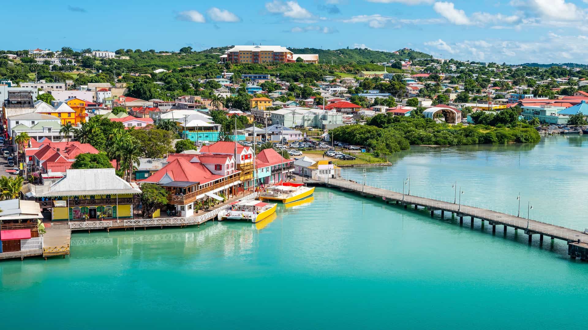 An aerial view of the colorful waterfront of St. John's, Antigua, with a long pier extending into the calm, blue sea and the town's charming buildings and marina in the background.