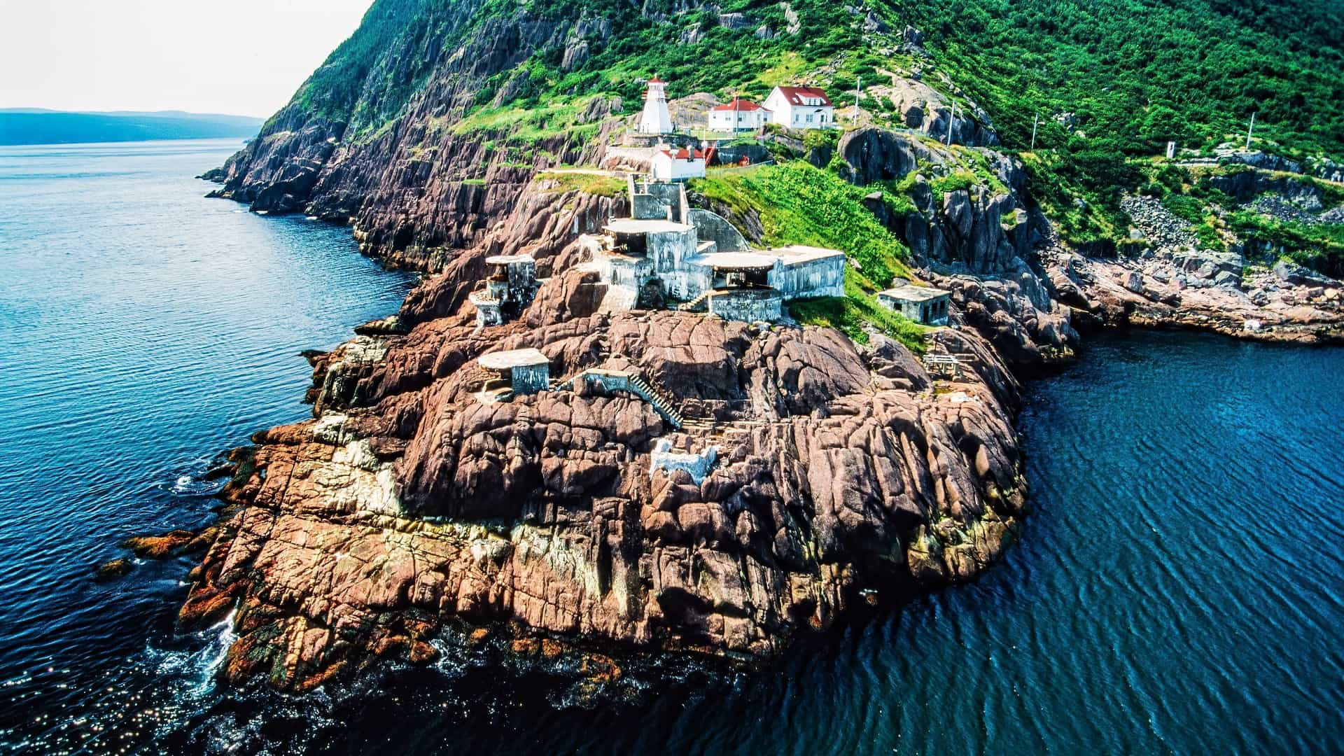 An aerial view of the rocky coastline of St. John's, Newfoundland, with a long, brown rocky point with abandoned buildings and a lighthouse on a lush green cliff, jutting out into the deep blue sea.