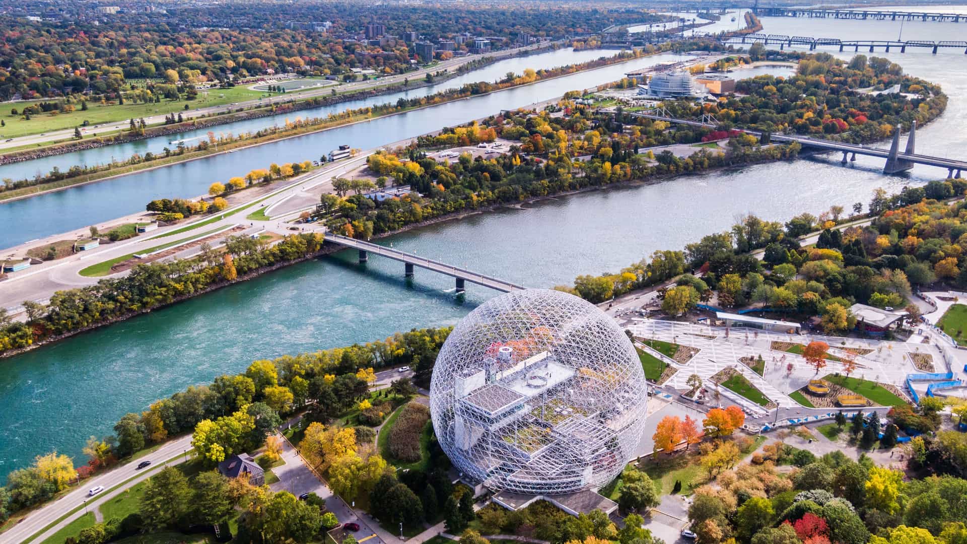 An aerial view of the Biosphere, a large geodesic dome, on an island in the St. Lawrence River in Quebec, Canada, surrounded by lush green and autumn-colored trees and several bridges.