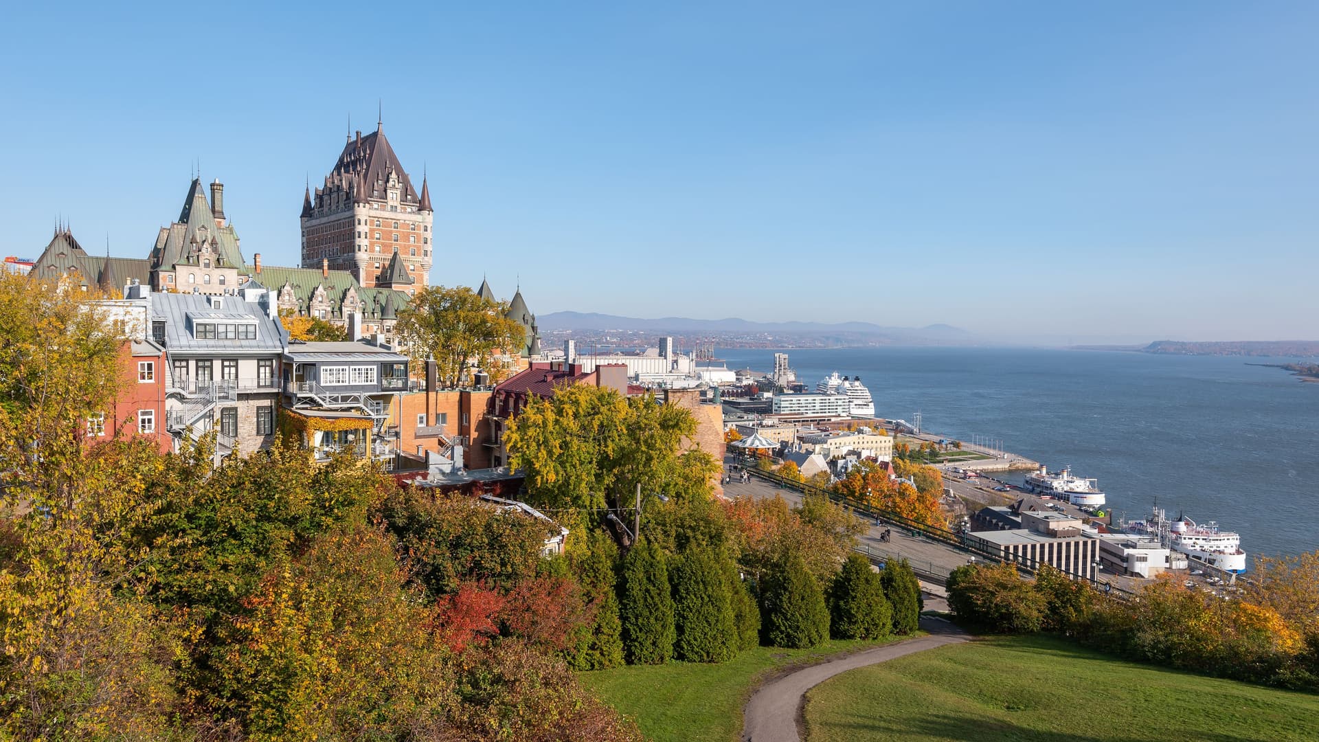 A high-angle view of the iconic Fairmont Le Château Frontenac hotel and the historic city of Quebec, with the St. Lawrence Seaway and a cruise ship in the background.