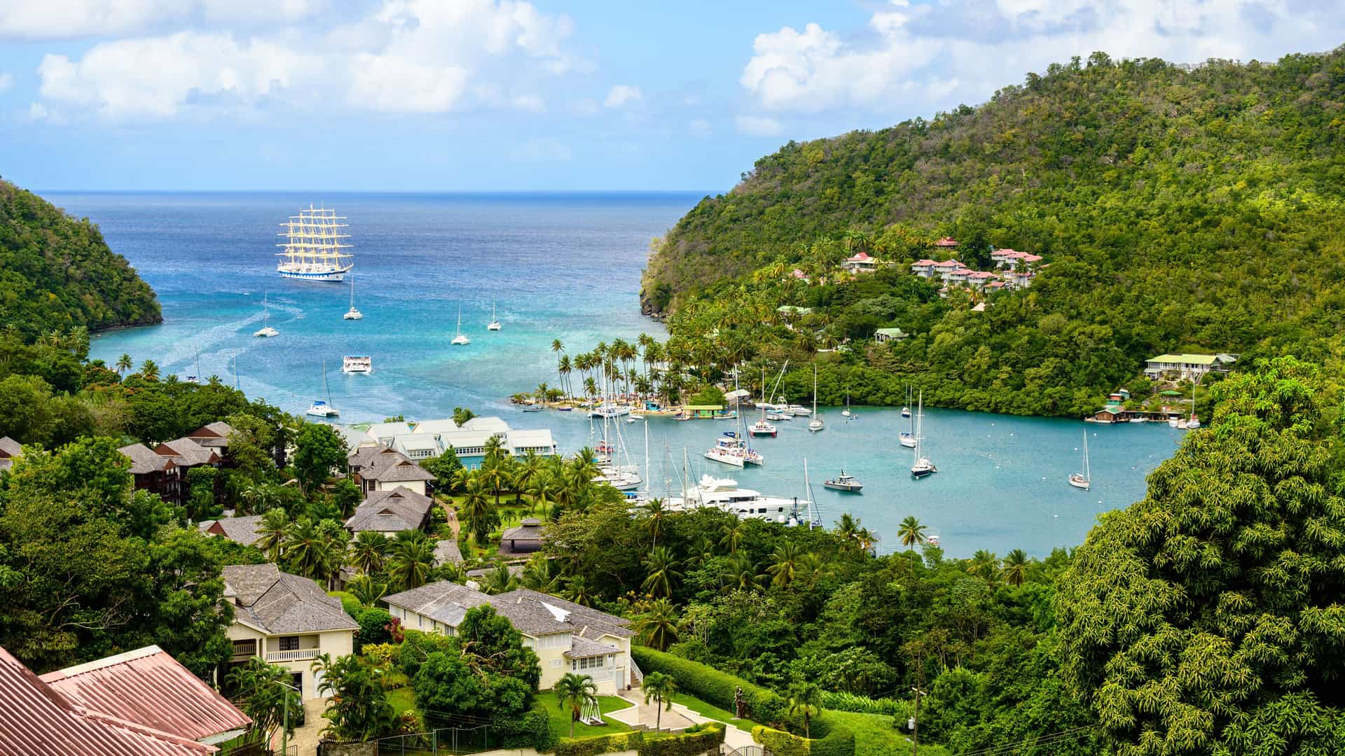 "A panoramic view of Marigot Bay, St. Lucia, with a tall-ship sailboat and other boats on the calm blue water, surrounded by lush green hills and tropical resorts. "