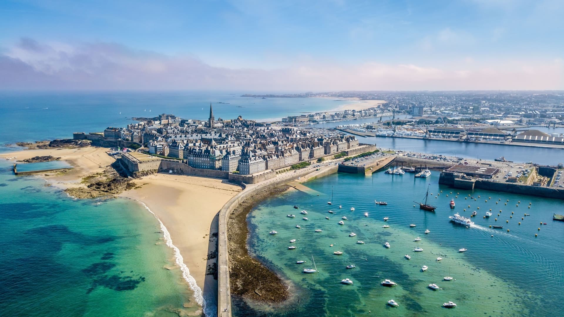 An aerial view of the historic walled city of St-Malo, France, with its medieval buildings and walls, a sandy beach, and a harbor full of sailboats and boats in the foreground.