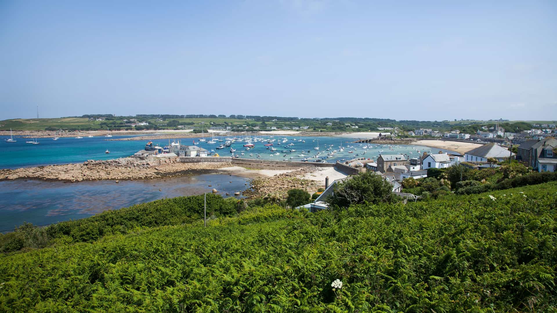 A wide-angle, high-angle view of the scenic harbor of St. Mary's, Isle of Scilly, with numerous sailboats and boats docked in the calm, turquoise waters and a town of white houses nestled at the base of a lush green hill.